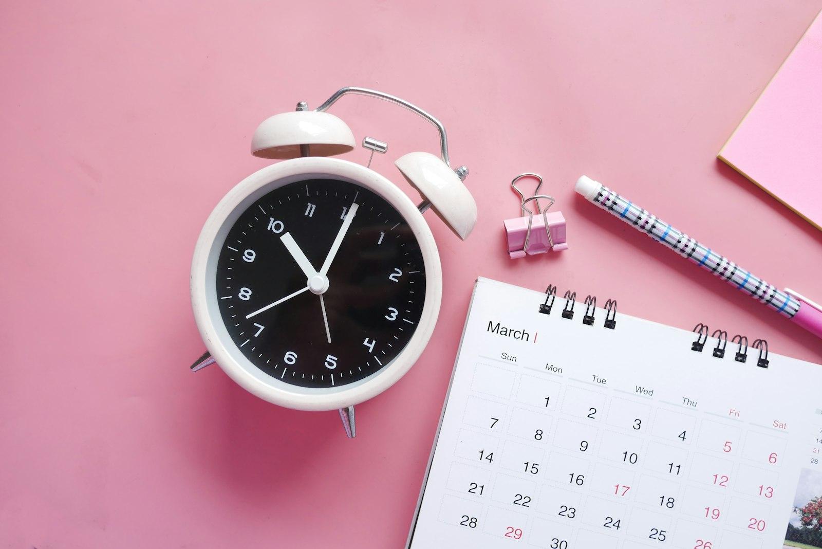 A white and pink analog alarm clock displayed with a calendar on a pink background