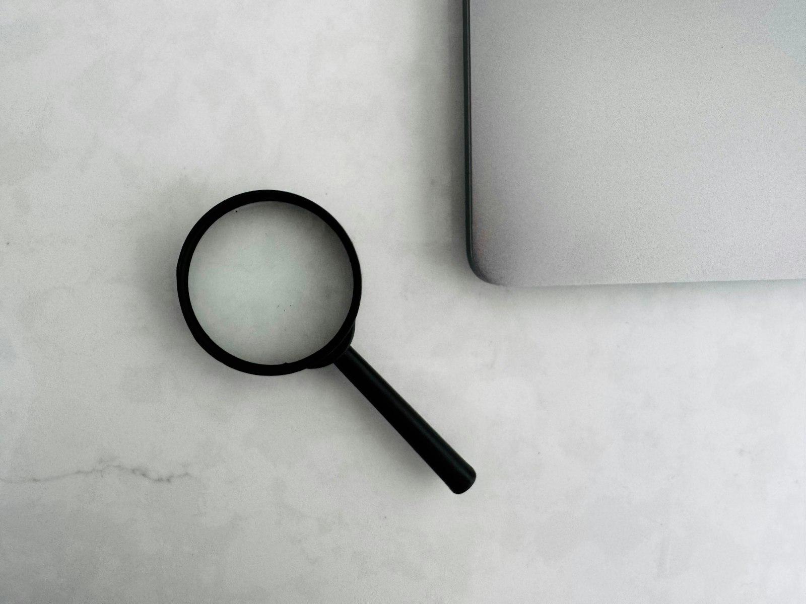 A black magnifying glass rests on a white marble countertop next to the corner of a closed silver laptop