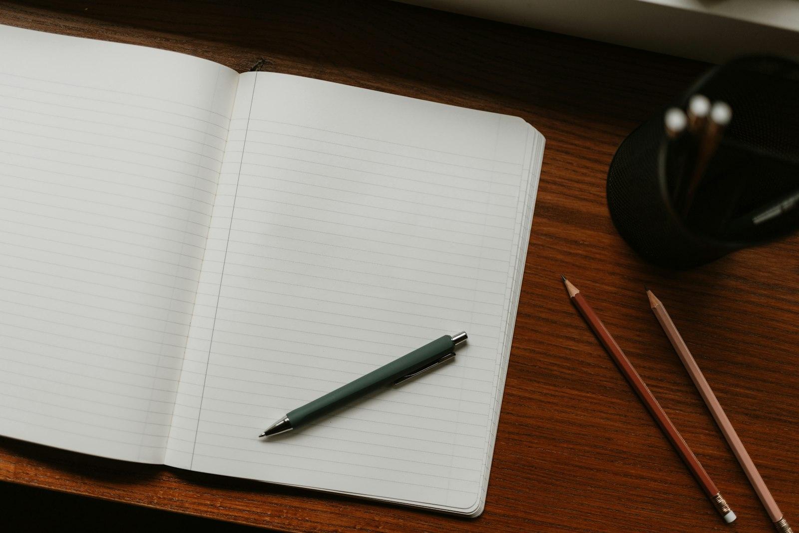 An open notebook with blank lined pages on a wooden desk, accompanied by green and purple writing implements