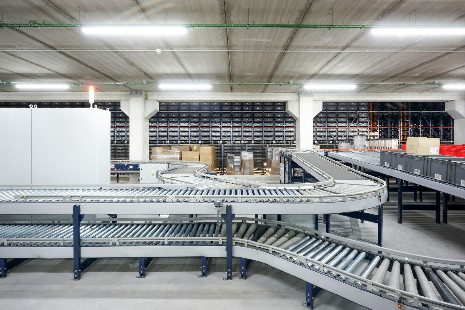 An empty industrial warehouse conveyor belt system curves between rows of storage racks under fluorescent lighting