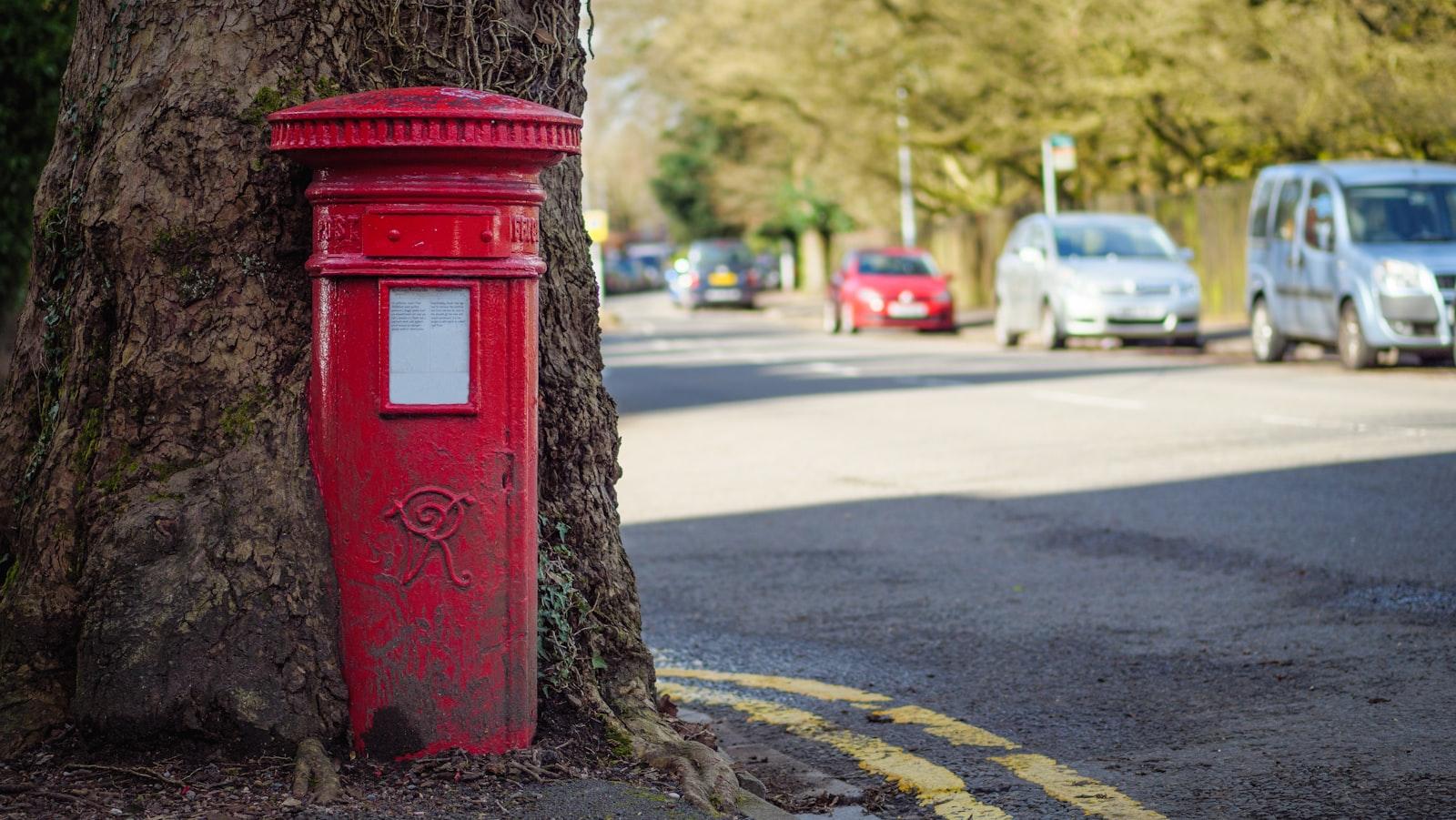 A red pillar-box style mailbox mounted against a tree trunk on a residential street, with cars parked along the road in the background