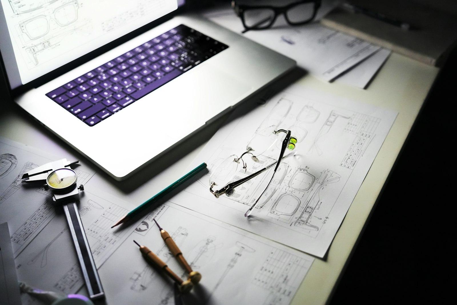 An open laptop sits on a desk surrounded by hand-drawn design sketches, blueprints, and drafting tools including a caliper and pencils