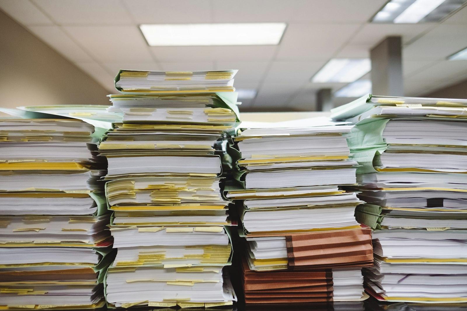 Stacked office papers and file folders depicting a busy workspace with numerous documents piled together