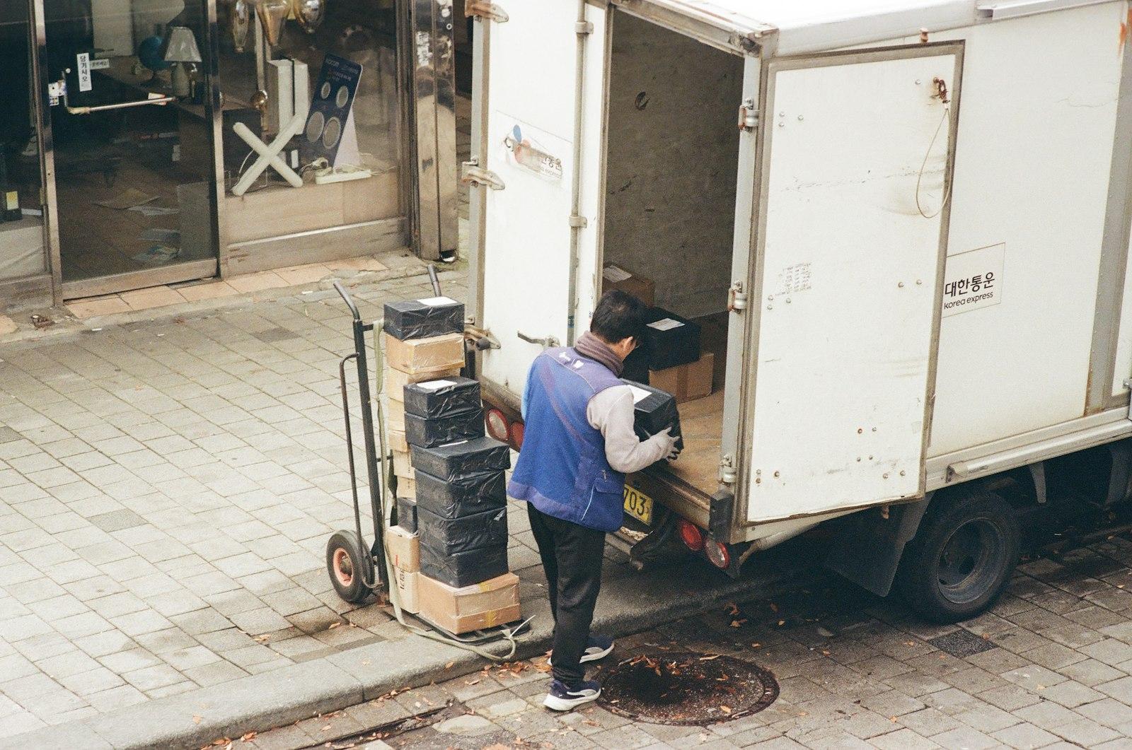 A delivery worker in a blue vest loads boxes from a hand truck into the open back of a white delivery van parked on a city sidewalk