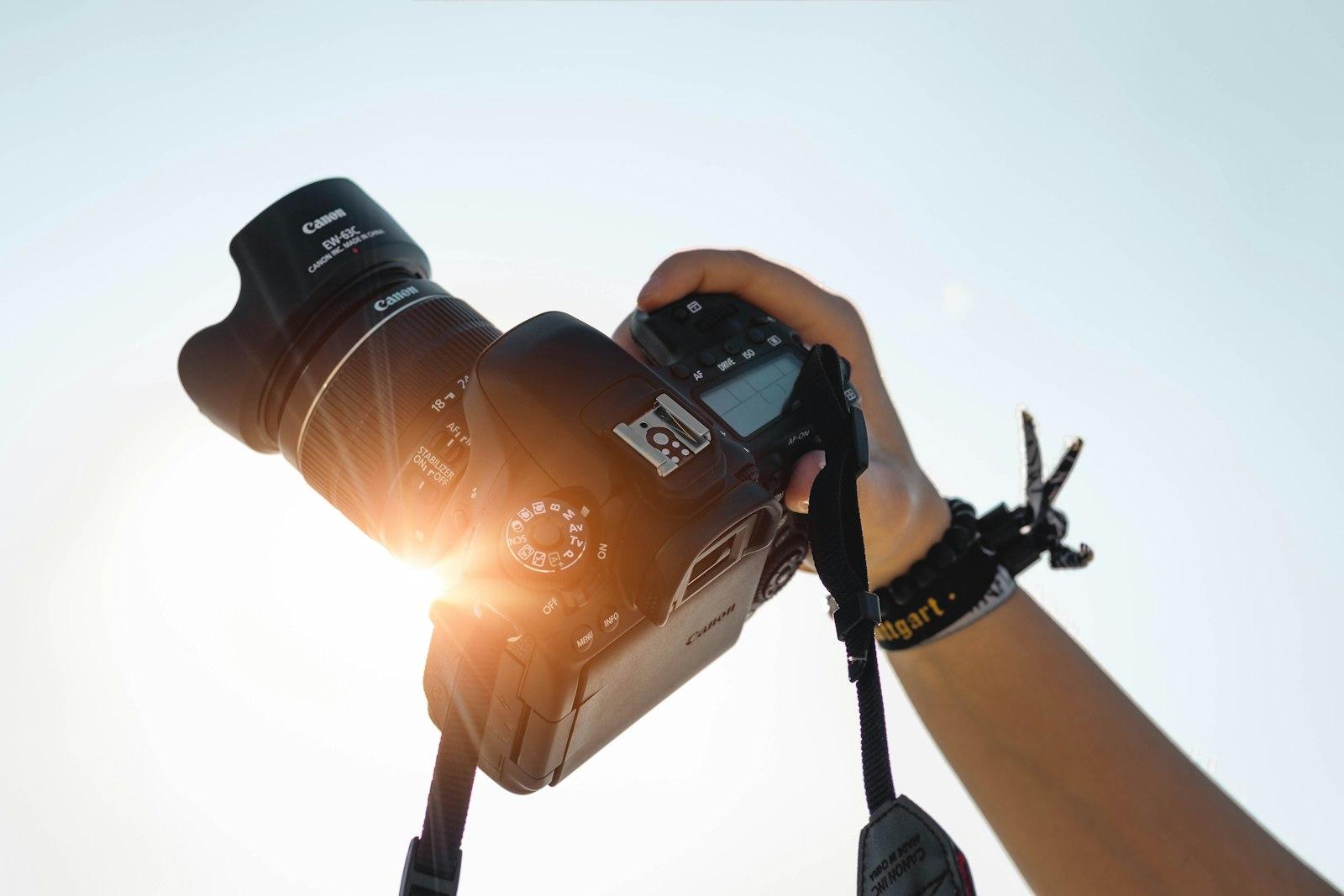 A hand holds up a Canon DSLR camera with a large zoom lens against a bright sky, sunlight flaring through the lens